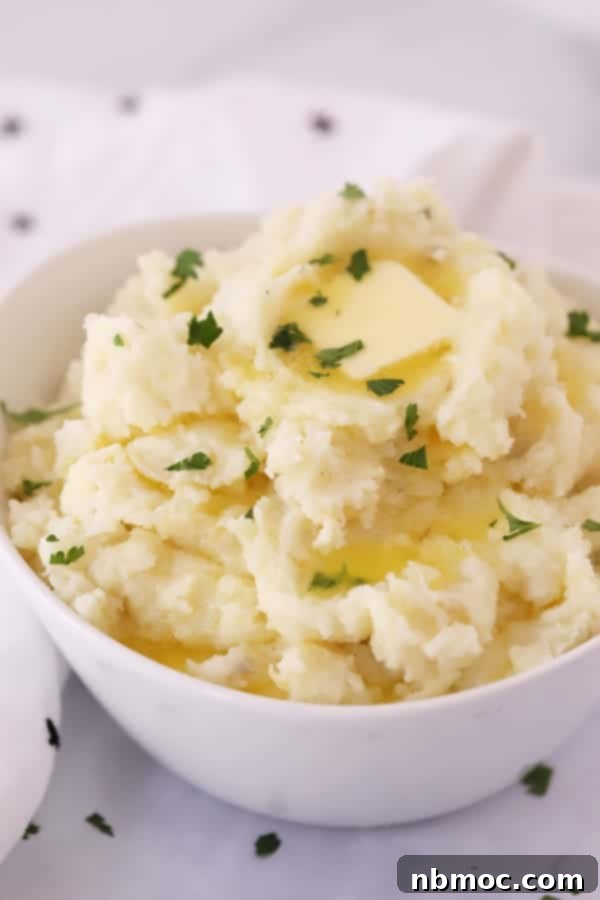A white serving bowl filled with creamy Gruyere mashed potatoes, garnished with a pat of melting butter and fresh chopped parsley.