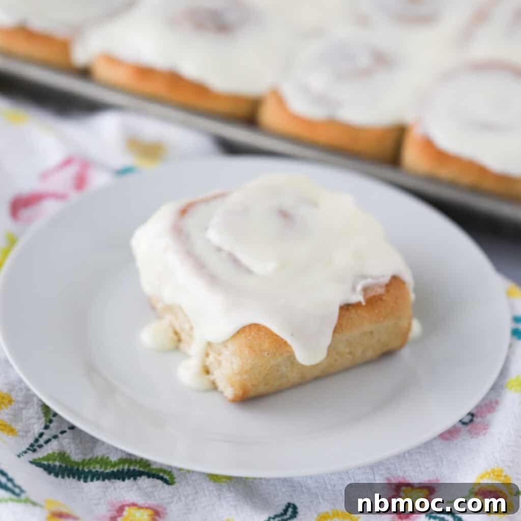 A perfectly baked cinnamon roll on a white plate, generously topped with creamy white cinnamon roll frosting, ready to be enjoyed.