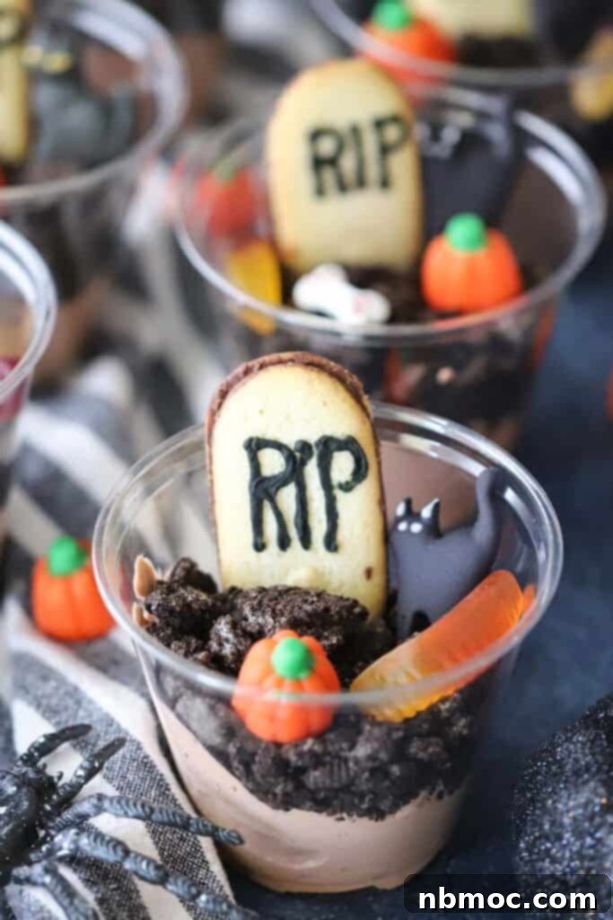 Haunted Earth Pudding Cups 2 Halloween pudding cups, decorated to look like graveyard pudding. Clear plastic cups show layers of chocolate pudding and dark Oreo crumbs, topped with Milano cookie gravestones, candy pumpkins, and gummy worms.