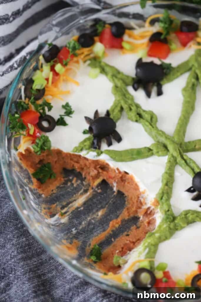 A large glass dish filled with vibrant Halloween Taco Dip, artfully decorated to resemble a spooky spider web with black olive spiders resting on top, ready for a party.