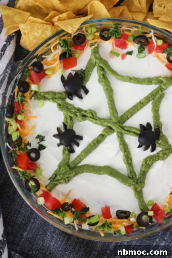 A close-up shot of the Halloween Bean Dip, showing the distinct layers and the perfectly piped guacamole spider web with olive spiders for decoration.