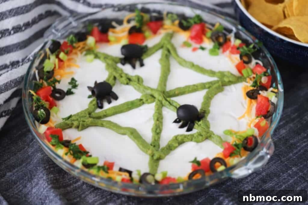 Halloween taco dip, showcasing its distinct layers and a perfectly crafted guacamole spider web with black olive spiders, garnished with fresh cilantro and lime wedges.