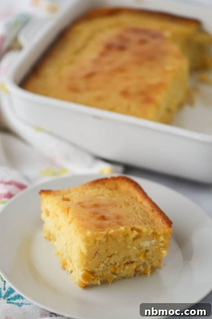 A slice of this creamed corn casserole recipe on a white serving plate with the full tray in the background, showcasing its golden-brown top and moist interior.