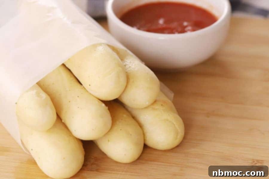 breadsticks olive garden wrapped in a sheet of parchment next to a small bowl of marinara dipping sauce, copycat olive garden breadsticks, best breadstick recipe.