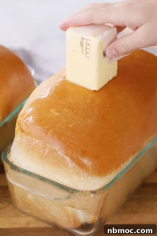 A loaf of freshly baked homemade white bread in a loaf pan with a hand spreading butter over the top, highlighting its golden crust and soft texture.
