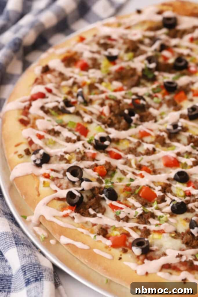 Fiesta Taco Pizza 2 A homemade taco pizza on a table topped with ground beef, olives, tomatoes, lettuce, and a mexican pizza sauce drizzle.
