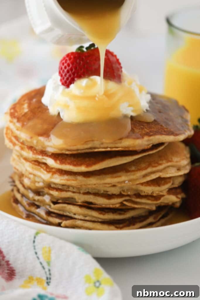 A tall stack of fluffy whole wheat pancakes with golden maple syrup being poured over the top, ready to be enjoyed.