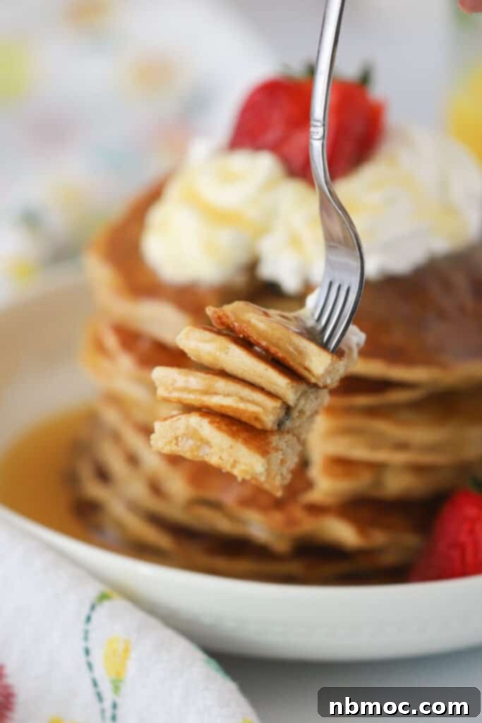 A fork holding a piece of a fluffy whole wheat pancake, topped with a dollop of whipped cream and a fresh strawberry slice.