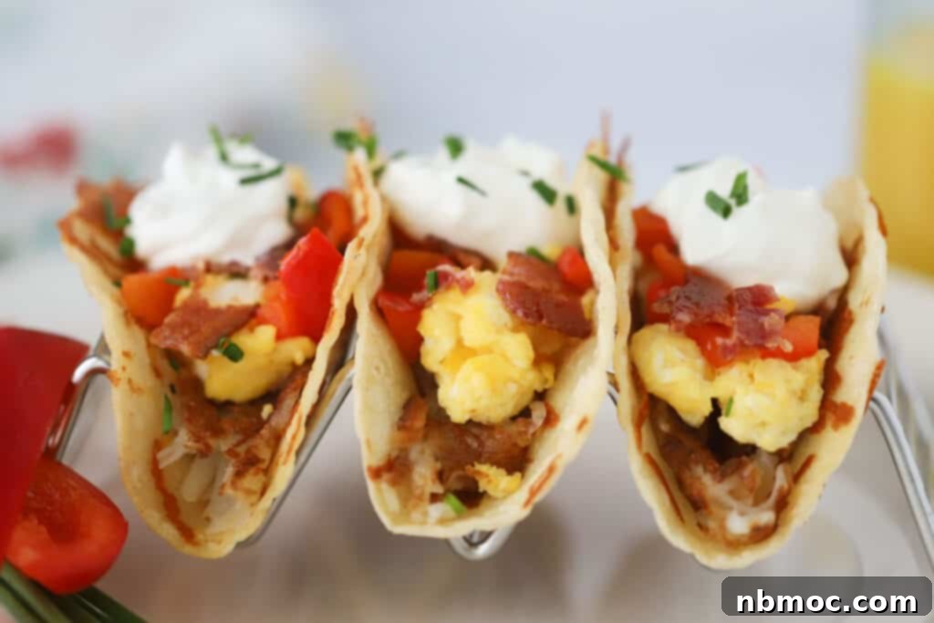 Easy breakfast tacos in a taco holder filled with hash browned potatoes, scrambled eggs, bacon bits, veggies, and topped with sour cream.