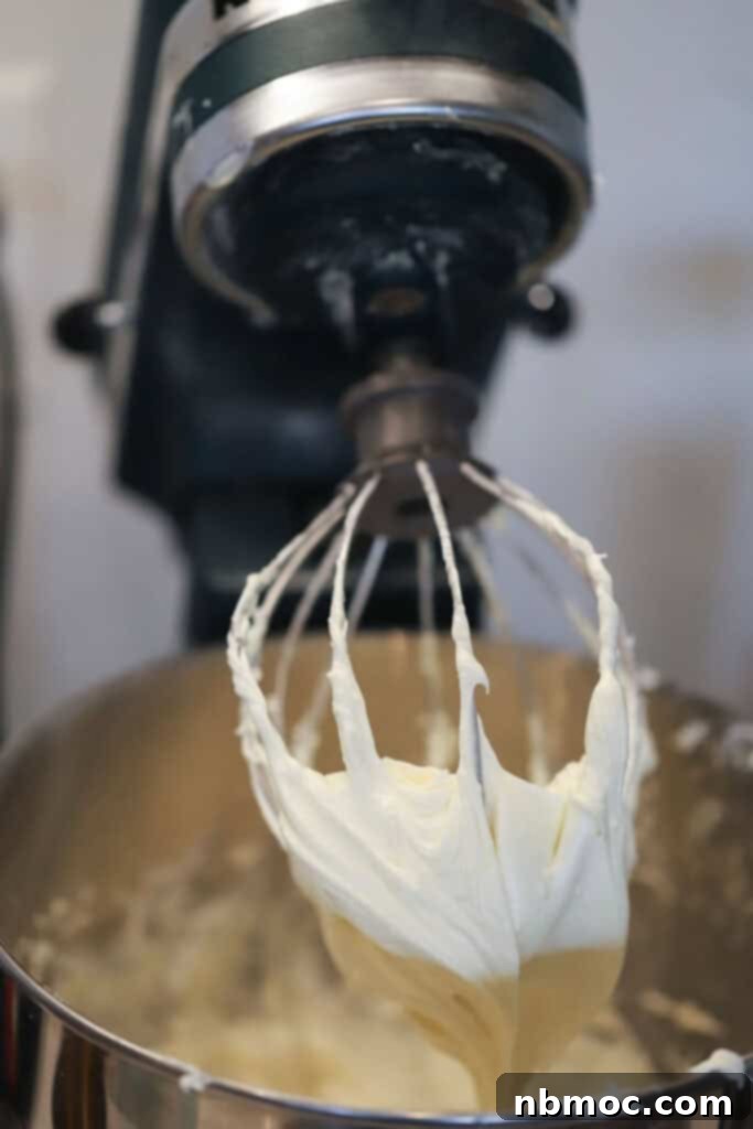 A stand mixer with the whisk attachment and a bowl full of white chocolate frosting.