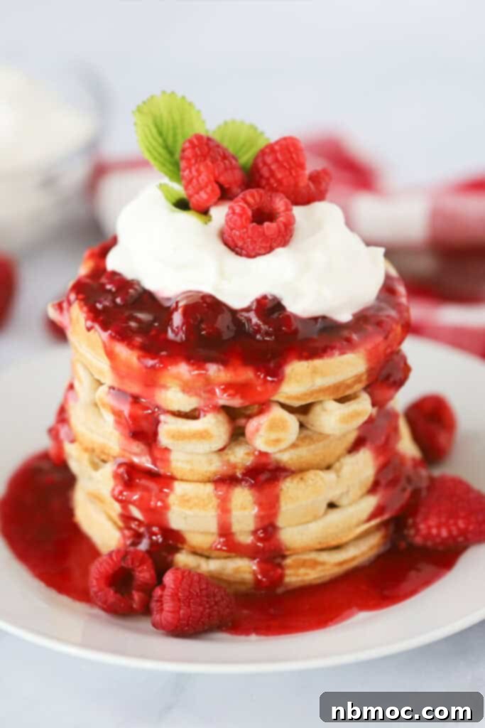 A stack of golden waffles generously topped with homemade, thick raspberry syrup and a swirl of whipped cream.