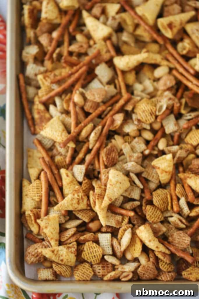 A baking sheet neatly covered with parchment paper, holding an even layer of seasoned Chex Party Mix, perfectly prepped for toasting in the oven.