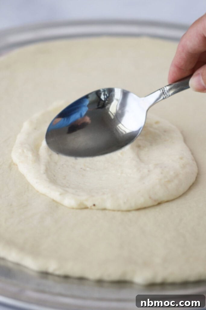 Creamy White Pizza Sauce 4 A spoon spreading this white sauce recipe for pizza over pizza dough.