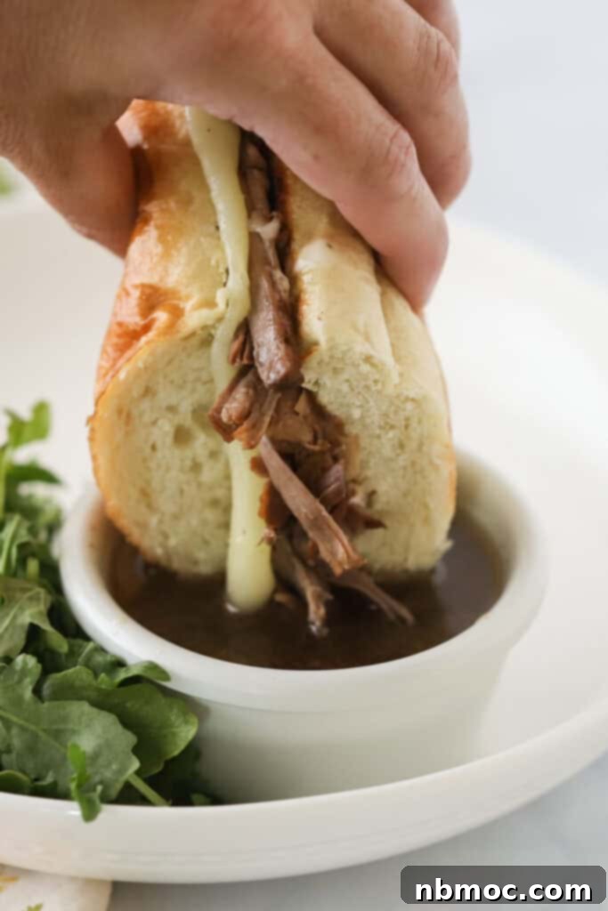 A hand dunking a slow cooker French Dip sandwich into au jus sauce, showcasing the juicy beef and melted cheese.
