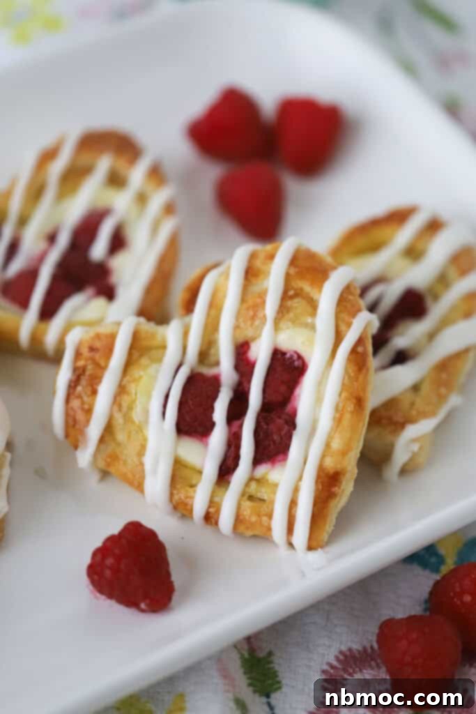 A white plate with a few raspberry danish filled with cream cheese and topped with glaze. These homemade pastries are made with fluffy puff pastry and fresh raspberries, perfect for breakfast.