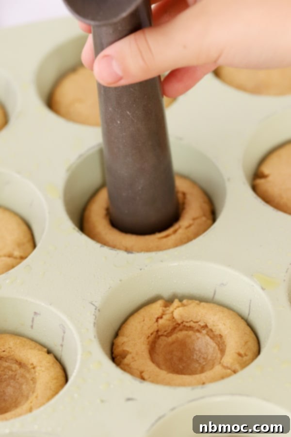 Peanut butter cookies in a muffin tin being pressed in the center with an ice cream scoop to make s'mores cookie cups.